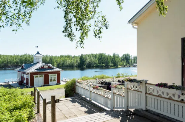 Kaunis näkymä järvelle, jossa punamultaista rakennusta ympäröi vihreä luonto ja kukkalaatikot.