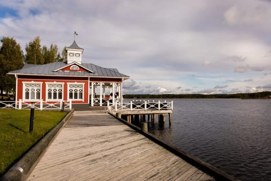 Punavalkoinen huvilarakennus laiturilla järven rannalla, taustalla kirkas sininen taivas ja levittäytyvät puut.