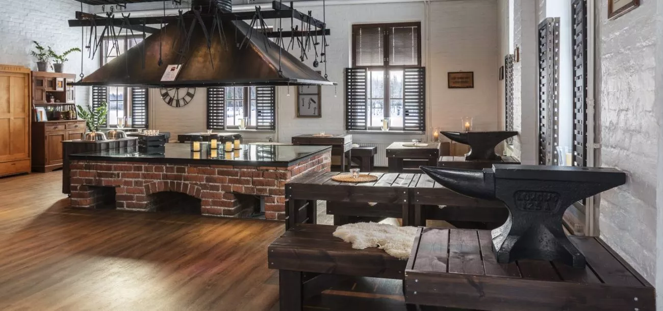 Moderni ravintola, jossa on tiiliseinä, suuret puiset pöydät ja industrial-designin kattovalaisin, sekä avokeittiö keskellä tilaa.