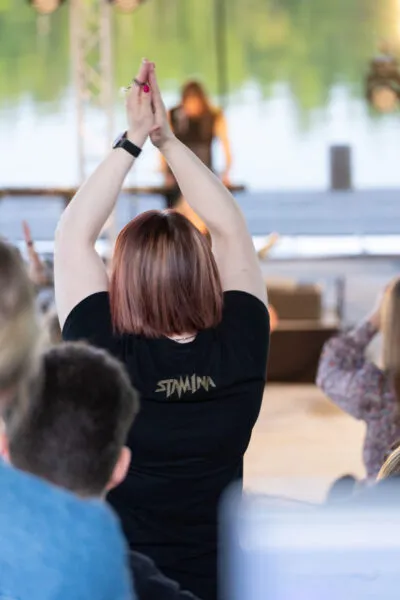 Nainen, joka seisoo käyttäen mustaa T-paitaa, taputtaa innokkaasti esitykselle järven rannalla tapahtuvassa konsertissa.