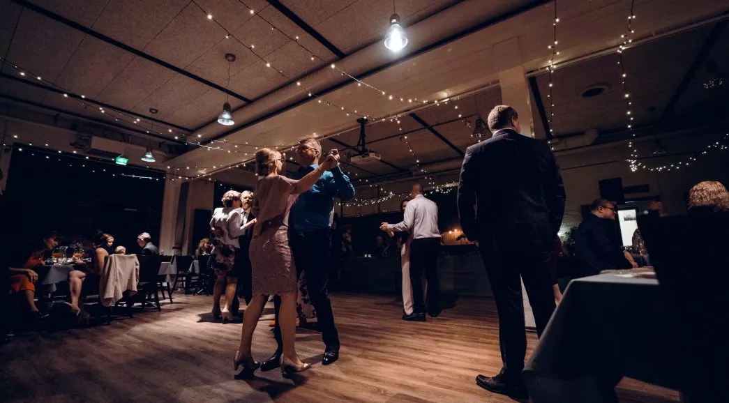 Kaksi paria tanssii romanttisessa juhlatilassa, jossa on string-valoja ja iloista tunnelmaa.
