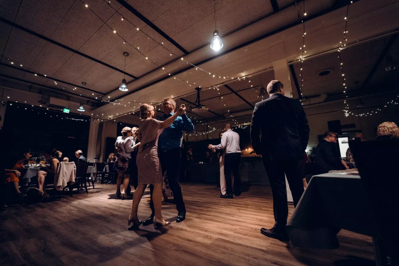 Kaksi paria tanssii romanttisessa juhlatilassa, jossa on string-valoja ja iloista tunnelmaa.