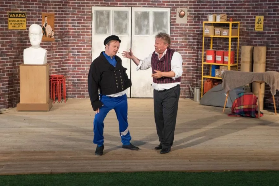 Kaksi miestä esittää teatterikohtausta lavalla, jossa on tiiliseinä taustalla ja valkoinen patsas.