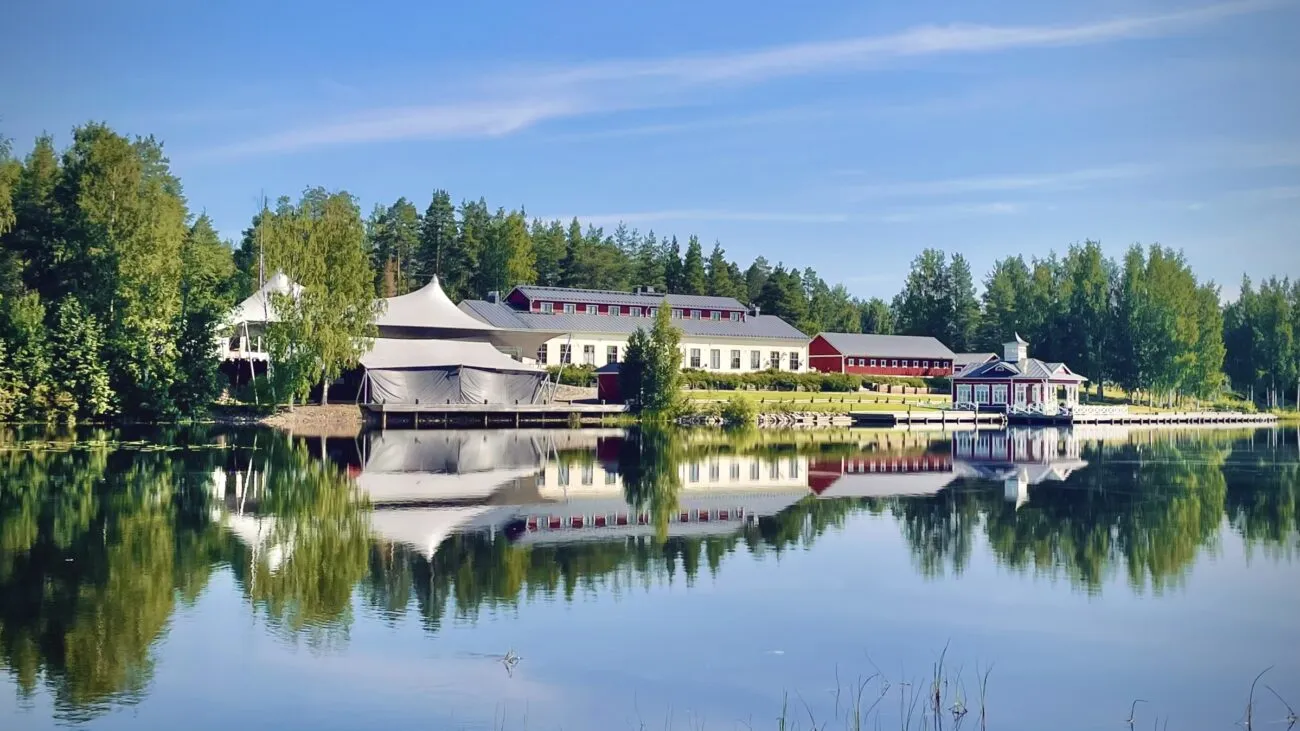 Viehättävä järvimaisema, jossa heijastuu valkoinen teltta ja punaiset sekä valkoiset rakennukset vehreiden puiden ympäröimänä.