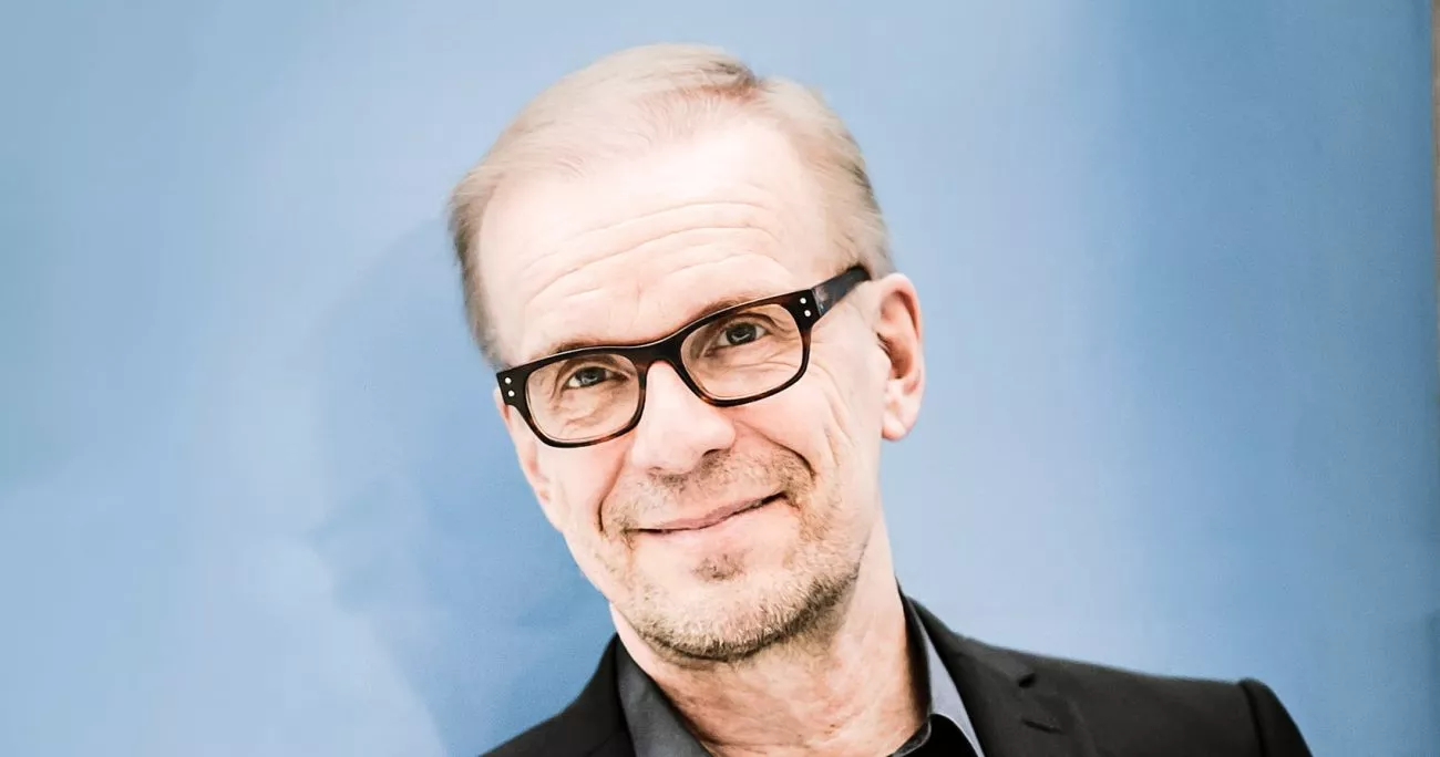 Hymyilevä keski-ikäinen mies kulmikkaissa silmälaseissa seisoo vaaleansinistä taustaa vasten.