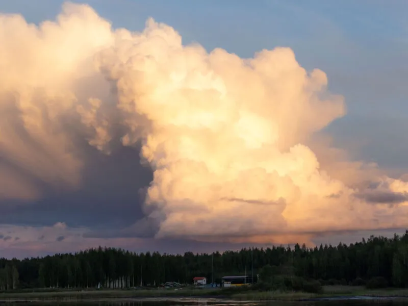 Pilvinen taivas hehkuu vaaleanpunaisena auringonlaskun aikaan, heijastuen rauhalliseen vesistöön, jonka äärellä on tiheä metsikkö ja muutama rakennus.