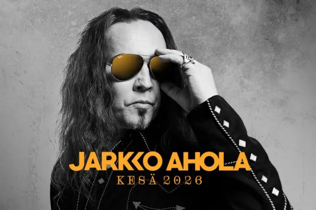 Jarkko Ahola, pitkät hiukset ja aurinkolasit, poseeraa tyylikkäässä mustassa takissa graafisessa taustassa.