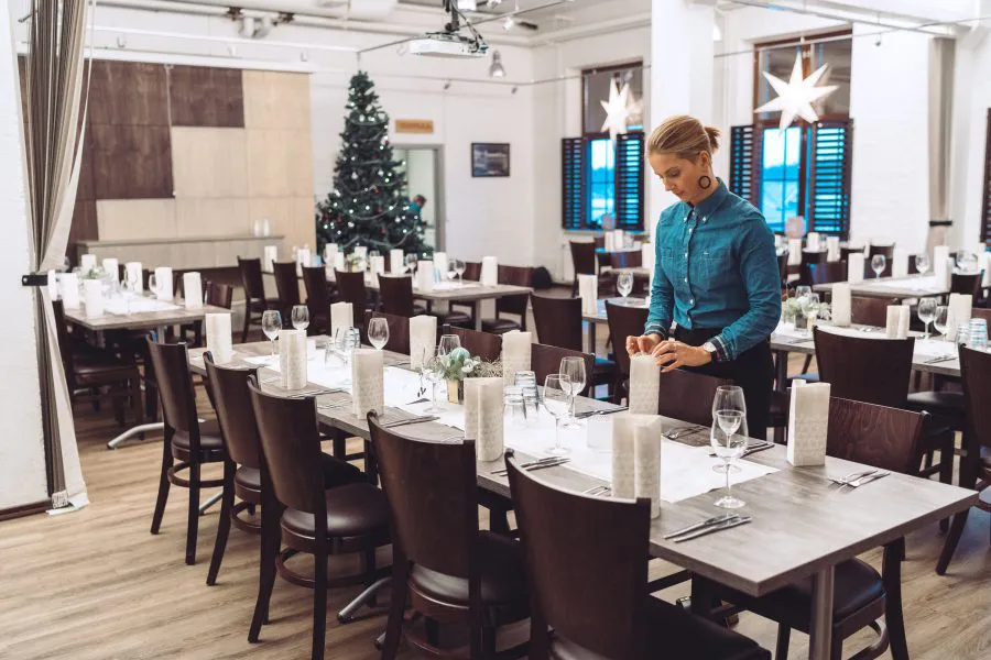 Jouluinen ravintola, jossa tarjoilija asettelee pöydät ja koristeet, taustalla joulukuusi.