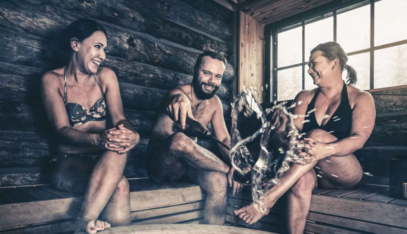Kolme henkilöä nauttii saunasta, heittävät vettä kiukaalle ja nauravat yhdessä.