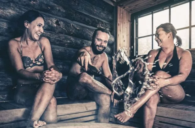 Kolme henkilöä nauttii saunasta, heittävät vettä kiukaalle ja nauravat yhdessä.