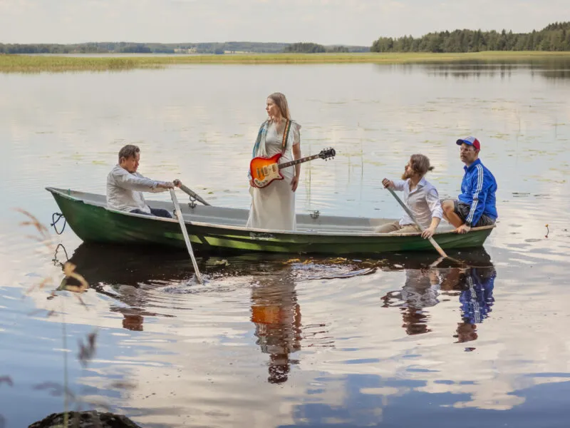 Neljä henkilöä veneessä järvellä, yksi soittaa kitaraa, kun muut nauttivat auringosta ja vesileikeistä.