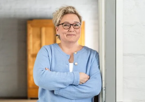 Nainen, jolla on vaaleansiniset vaatteet ja silmälasit, seisoo kädet ristissä hymyillen valkoisen seinän vieressä.