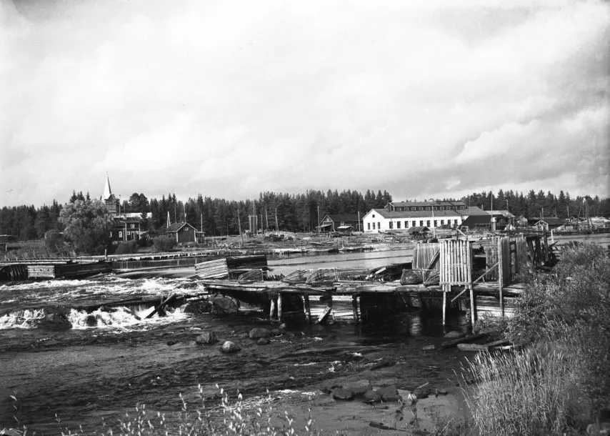 Vanha mustavalkoinen kuva koskesta, jossa on hirsirakenteinen rakennus ja metsää taustalla.