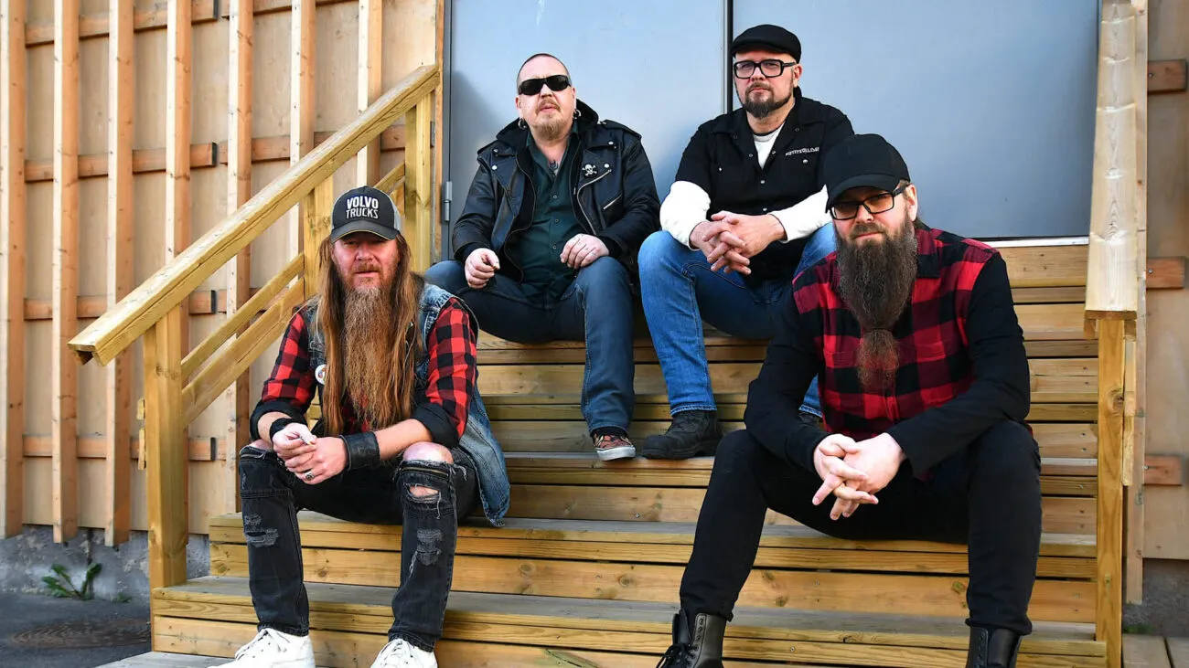 Neljä miestä istuu puutikkailla, pukeutuneina rock-tyyliin mustissa ja punaisissa vaatteissa.