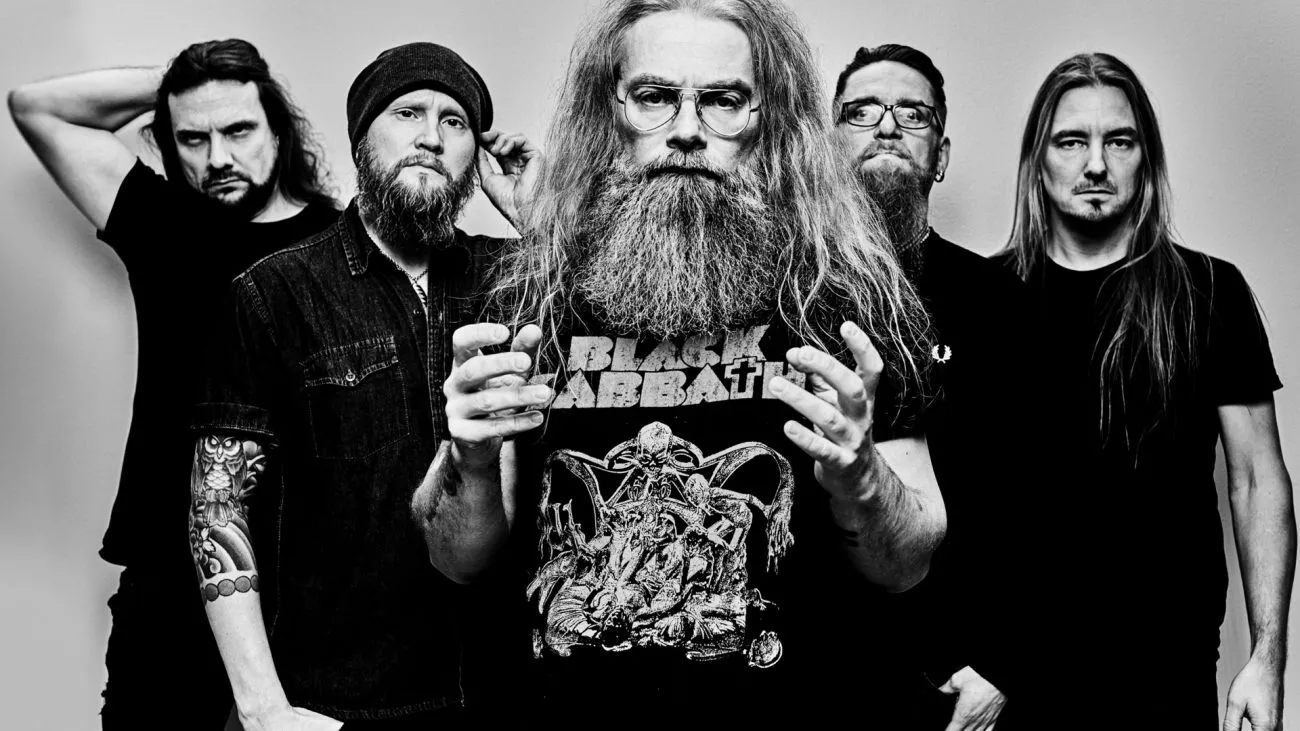 Viisi miestä seisoo ryhmässä, mukana pitkäpartainen ja -hiuksinen henkilö, joka pitää mustaa Black Sabbath -t-paitaa päällään.
