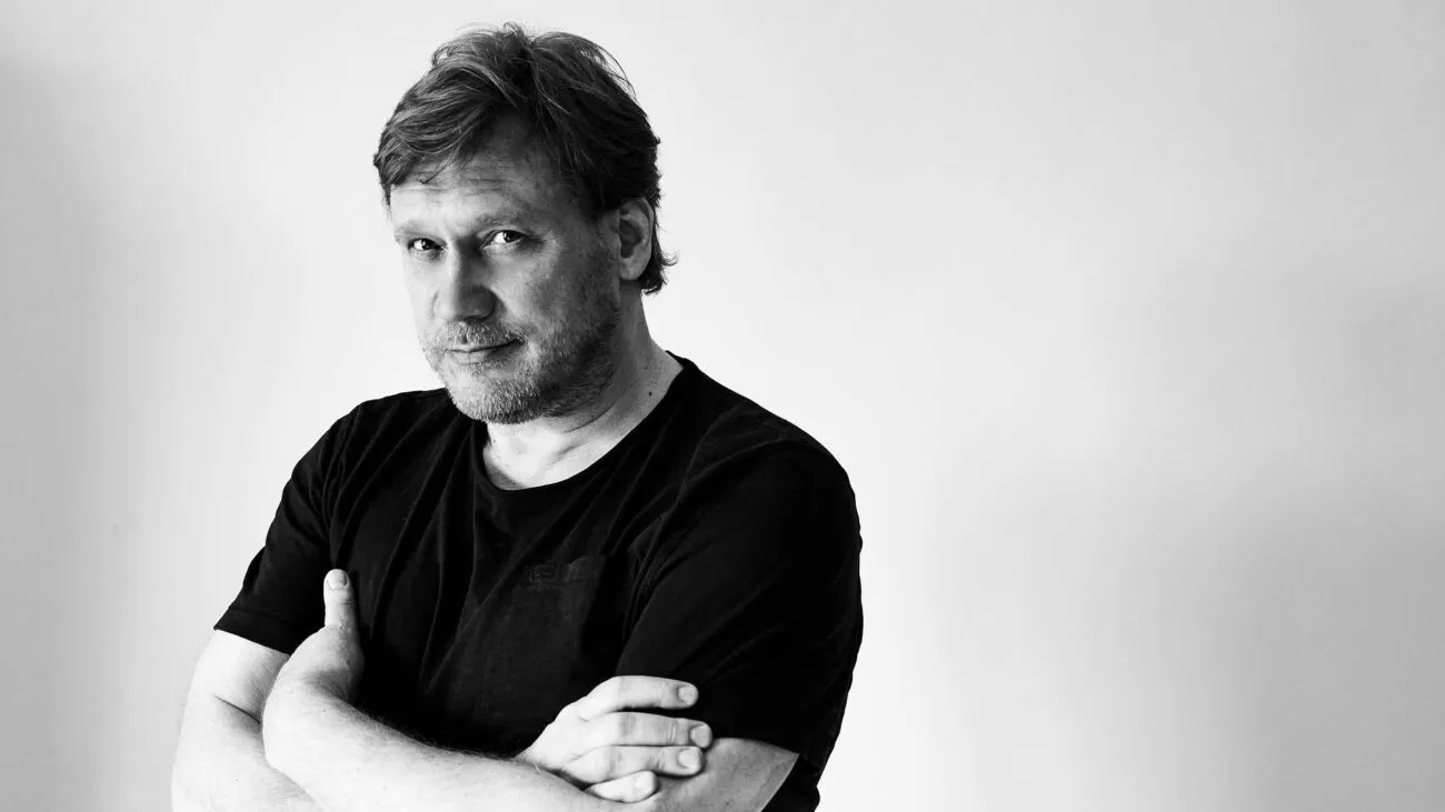 Mies, jolla on vaalea tukka ja parta, katsoo vakavasti kameraan mustassa t-paidassa, kädet ristissä rinnan kohdalla.