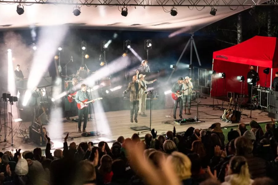 Energinen konsertti, jossa esiintyjät laulavat ja soittavat instrumentteja estradilla kirkastuvien valojen alla, ja yleisö nauttii esityksestä.