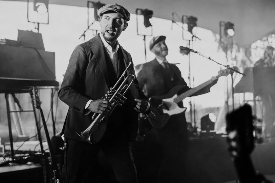 Muskettipäisessä musiikin esityksessä trumpettia soittava muusikko esiintyy lavalla, taustalla näkyy basisti ja valot.