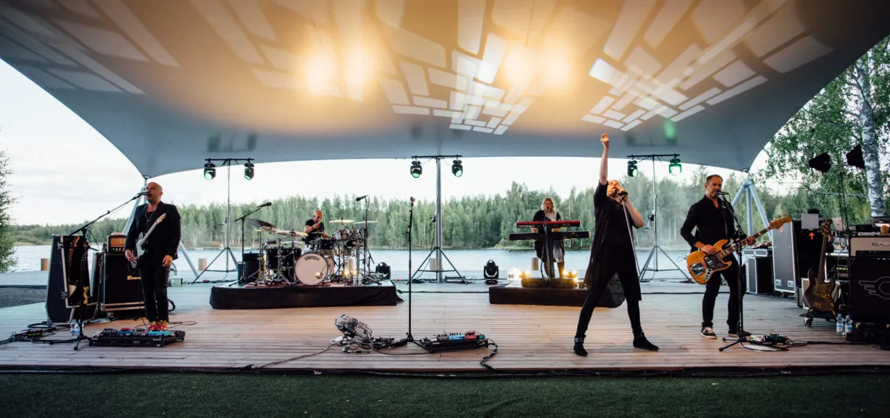 Rokkibändi esiintyy ulkoilmakonsertissa järven rannalla, valaistuksen luodessa näyttäviä varjoja katoksi.
