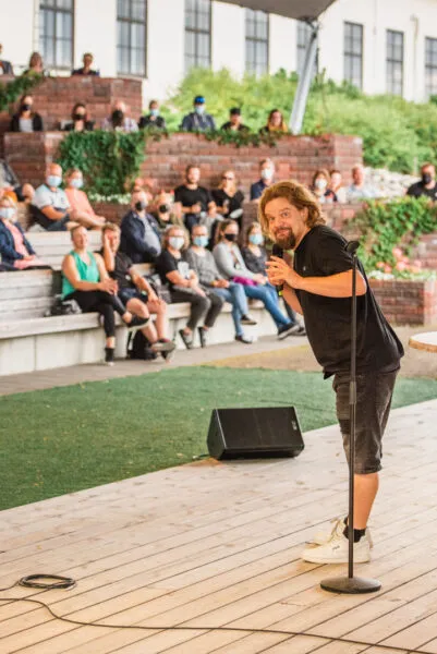 Kuvassa stand-up koomikko esiintyy ulkoilmatilassa yleisölle, joka istuu penkeillä ja keskittyy esitykseen.