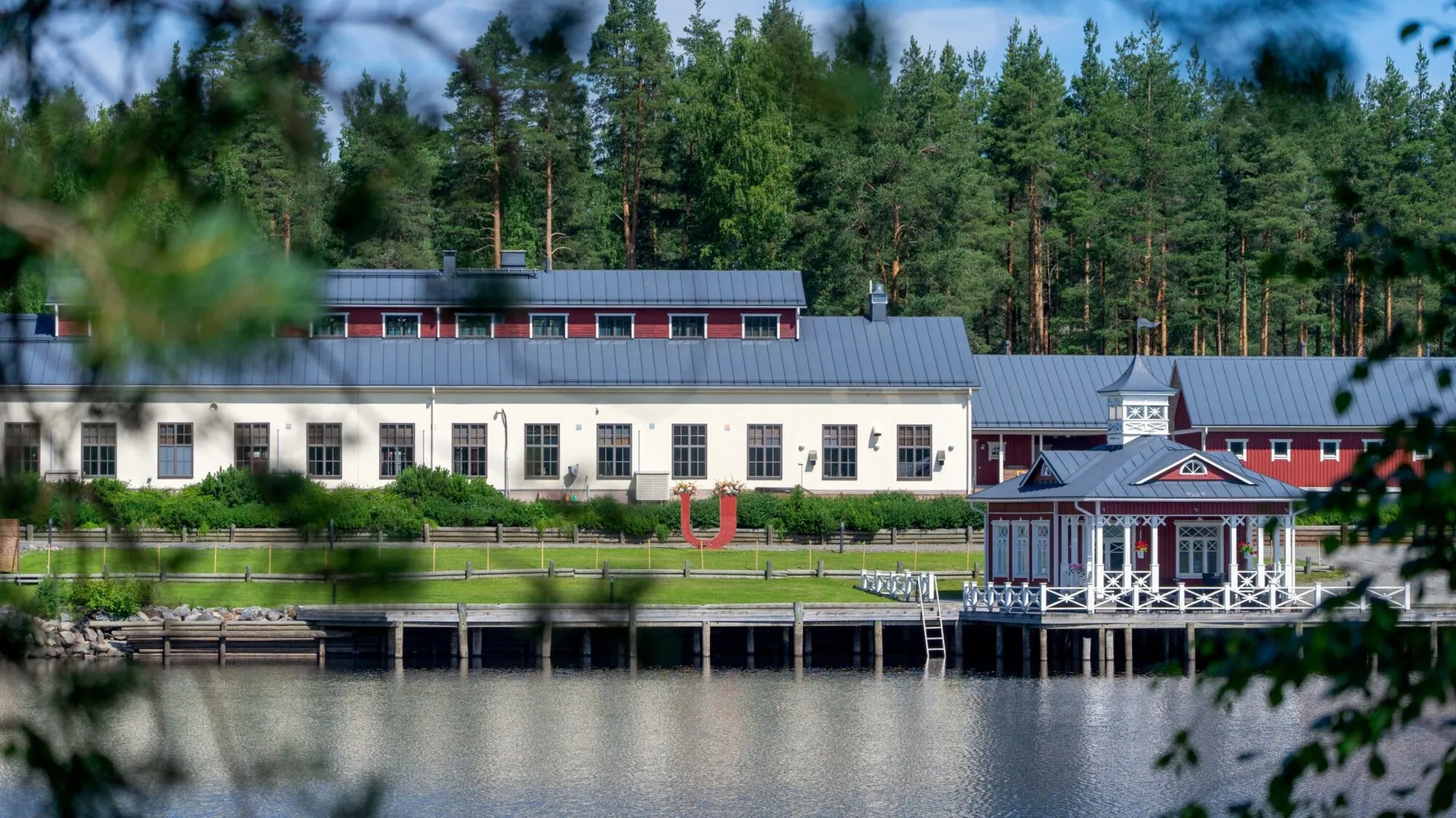 Kaunis jokirantamaisema, jossa näkyy Utran Uittotuvan päärakennus, paviljonki ja laituri vehreiden puiden takaa.