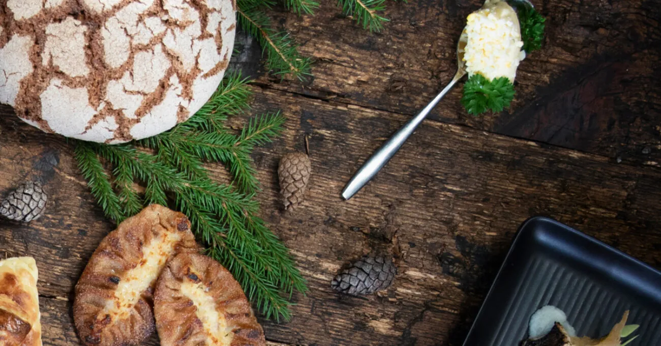 Ruisleipä, karjalanpiirakat, koristeltu ruokalusikka ja joulukuusen oksia puupinnalla.