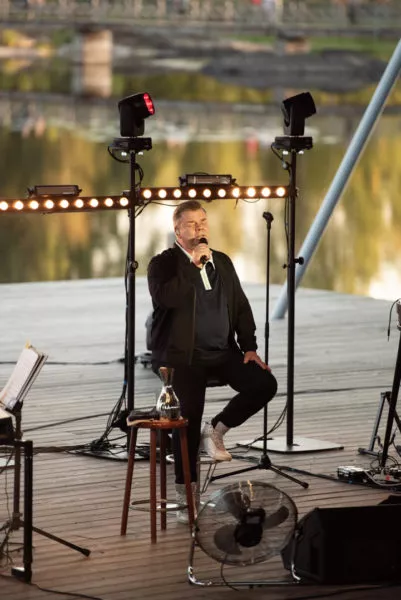 Mies istuu tuolilla lavalla, puhujamikrofoni kädessään, taustalla valaistuja valoja ja järvimaisema.