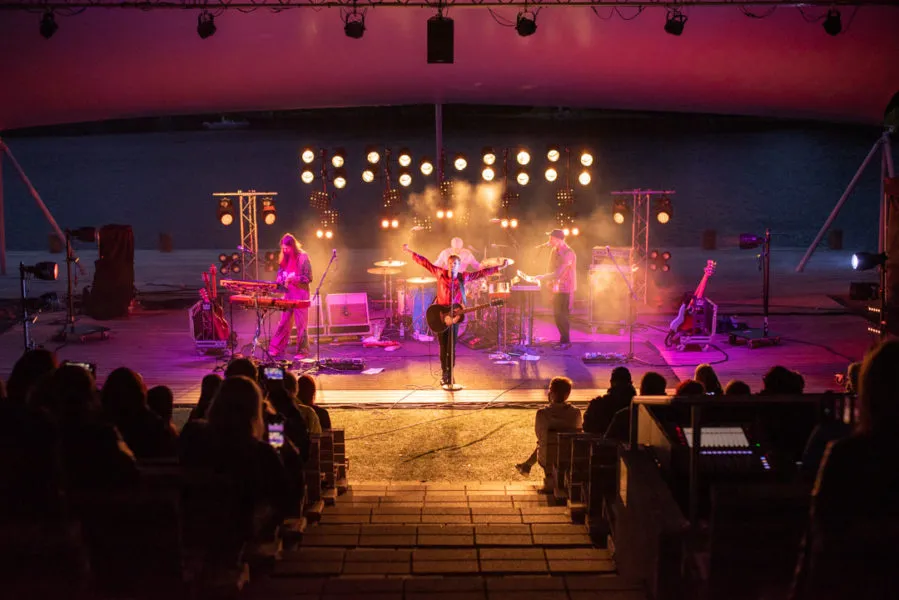 Muusikot esittävät konserttia ulkoilmalavalla, jossa on koristeellisia valoja ja katsojia istumassa eturivissä.