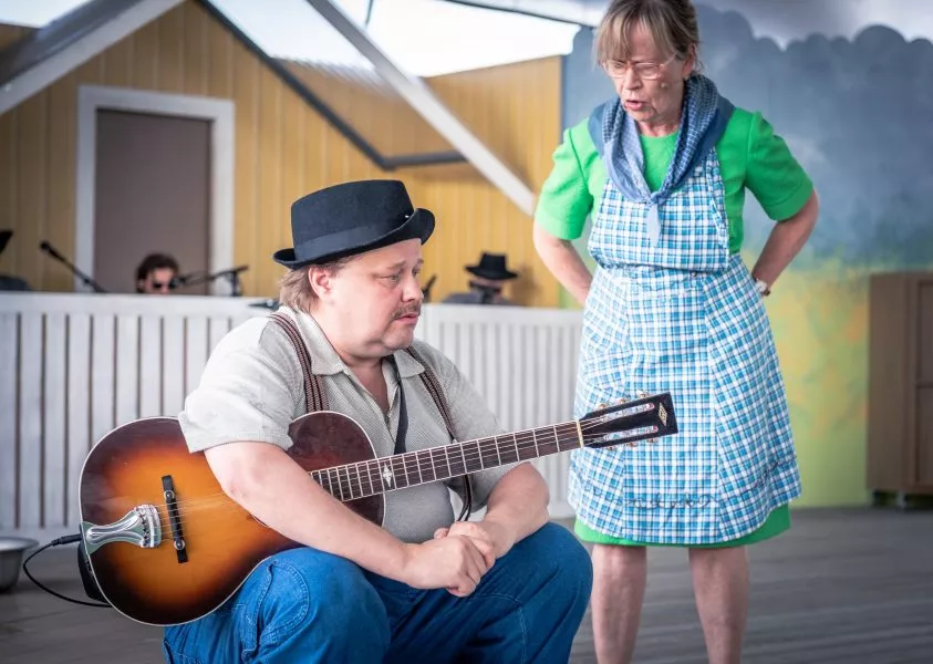 Mies istuu lattialla akustinen kitara sylissään, kun nainen puhuu hänelle vakavasti teatterilavalla.